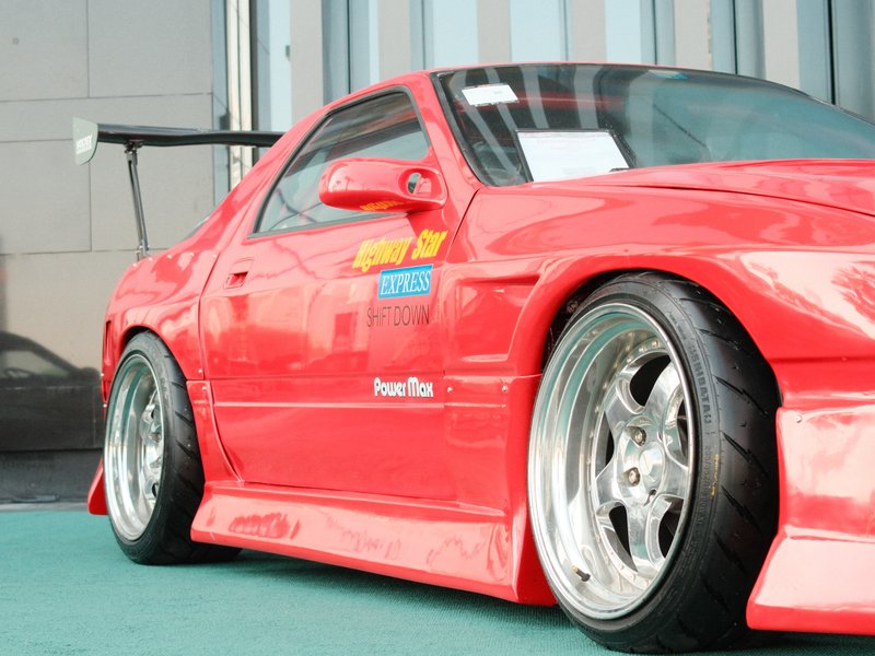 A red modified sports car with a large rear wing and shiny alloy wheels parked on a green surface.