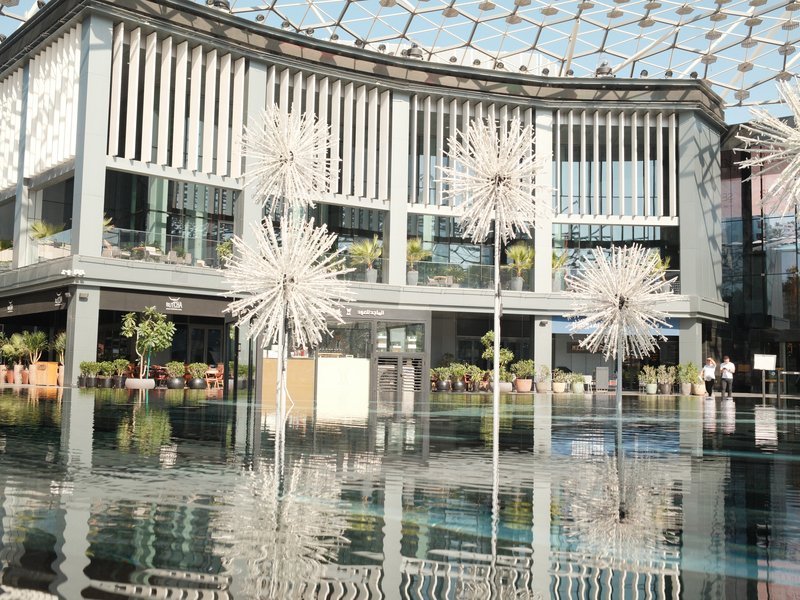 Modern indoor space with reflective water and artistic dandelion sculptures under a glass ceiling.