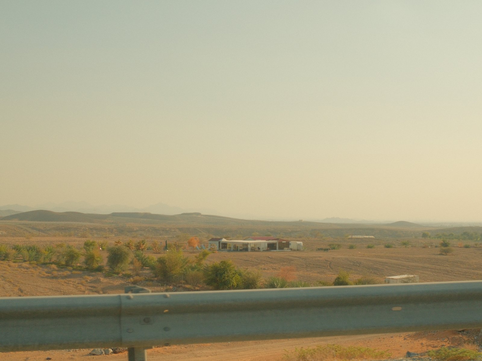 A distant view of a rural landscape with mountains and a building, seen over a highway guardrail.