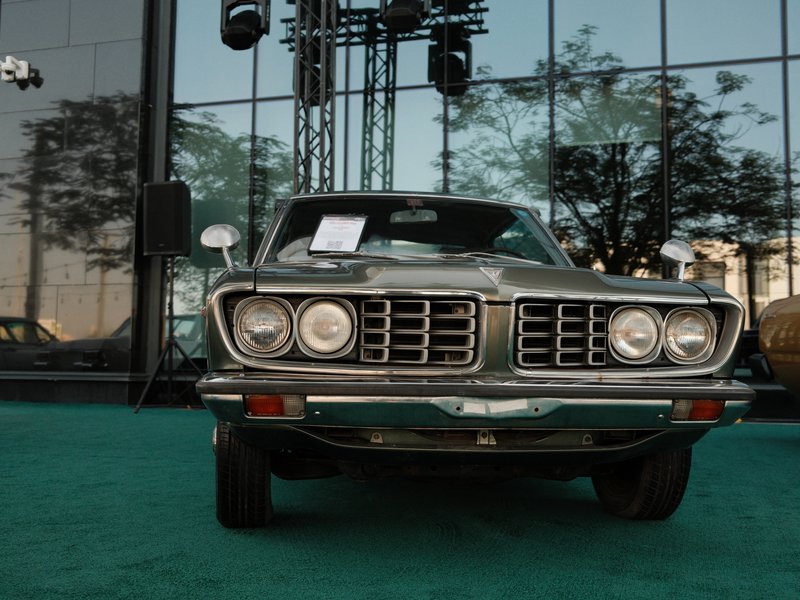 Front view of a vintage car parked in front of a glass building with reflections and greenery.