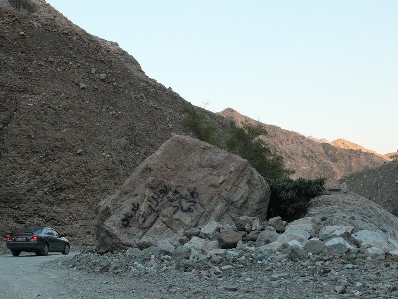 A rocky landscape with a large boulder and a car parked nearby, surrounded by mountains.