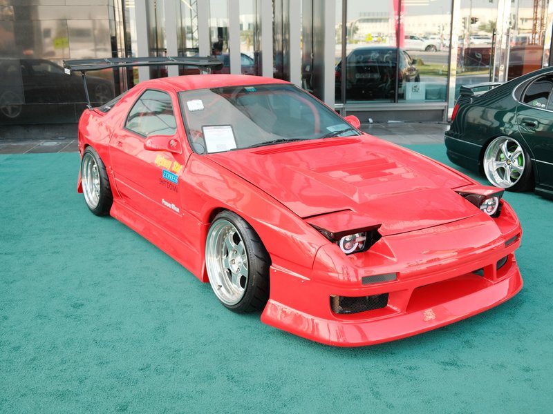 A bright red modified sports car with unique headlights parked on a teal carpet at an automotive event.