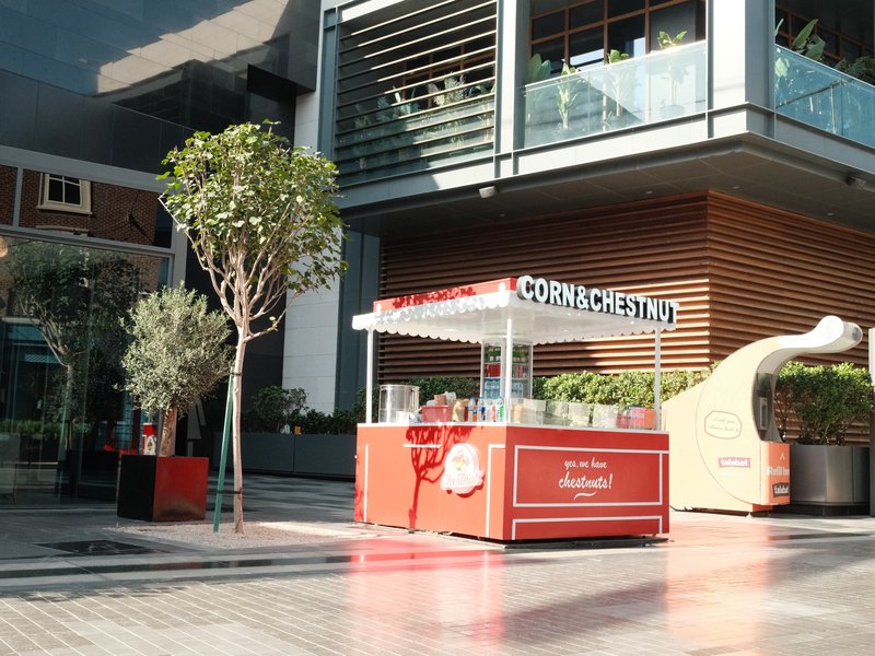 A bright outdoor scene featuring a food cart with a red canopy and nearby trees in an urban setting.