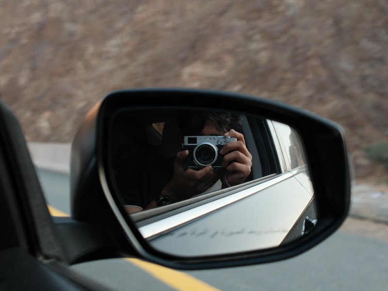A car side mirror reflecting a person holding a camera while driving on a road.