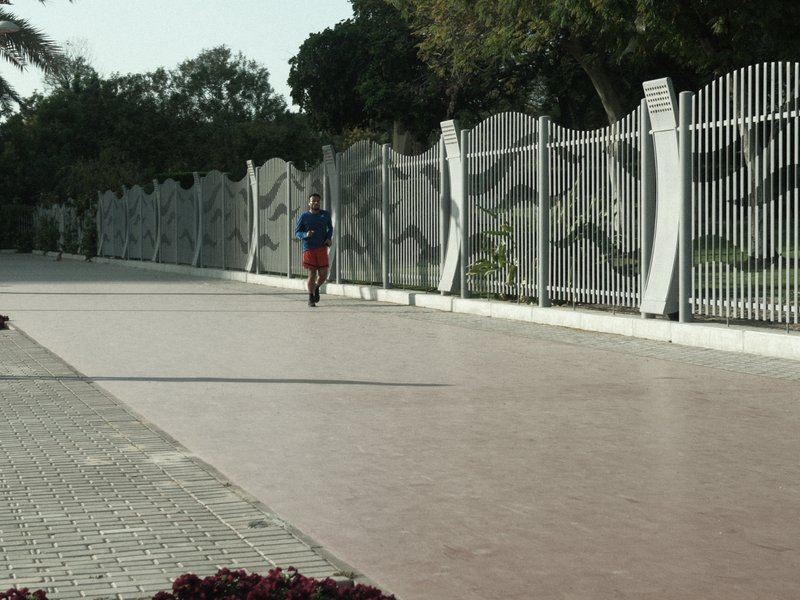A person riding a scooter on a wide, empty pathway lined with a decorative fence and greenery.