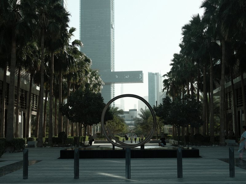 A modern urban plaza lined with palm trees and a circular sculpture, with skyscrapers in the background.