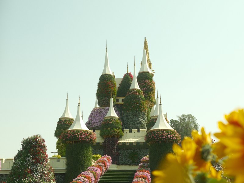 A whimsical castle adorned with colorful flowers against a clear sky, with sunflowers in the foreground.