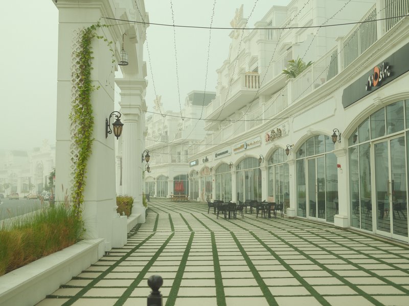 A misty outdoor walkway lined with white arches and greenery, leading to a series of shops.