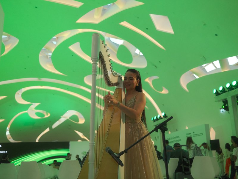 A woman playing a harp under a green-lit ceiling with abstract designs in a modern indoor setting.