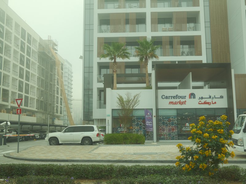 A modern building with a Carrefour store, palm trees, and a white car parked outside in a foggy atmosphere.