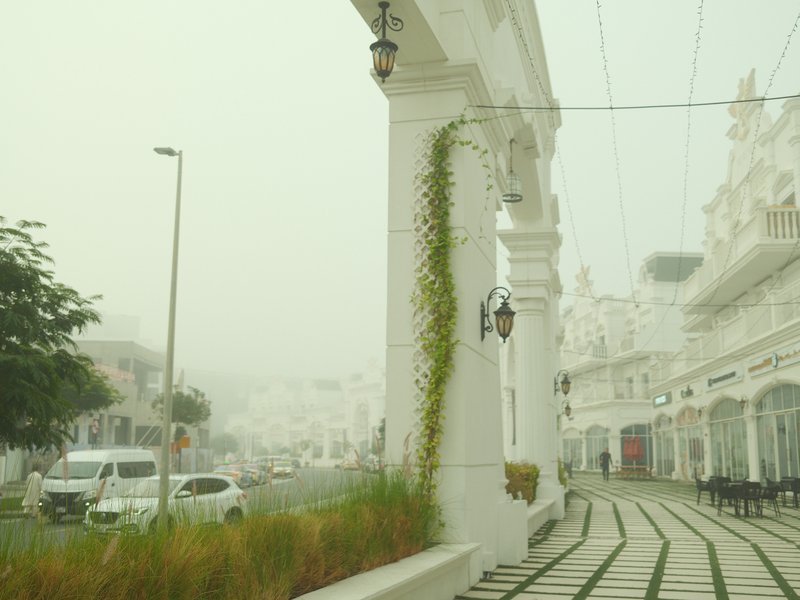 A foggy street scene with white buildings and greenery, featuring a decorative archway.