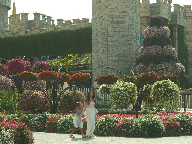 A woman in a white dress stands with a child in a vibrant flower garden near a stone castle.
