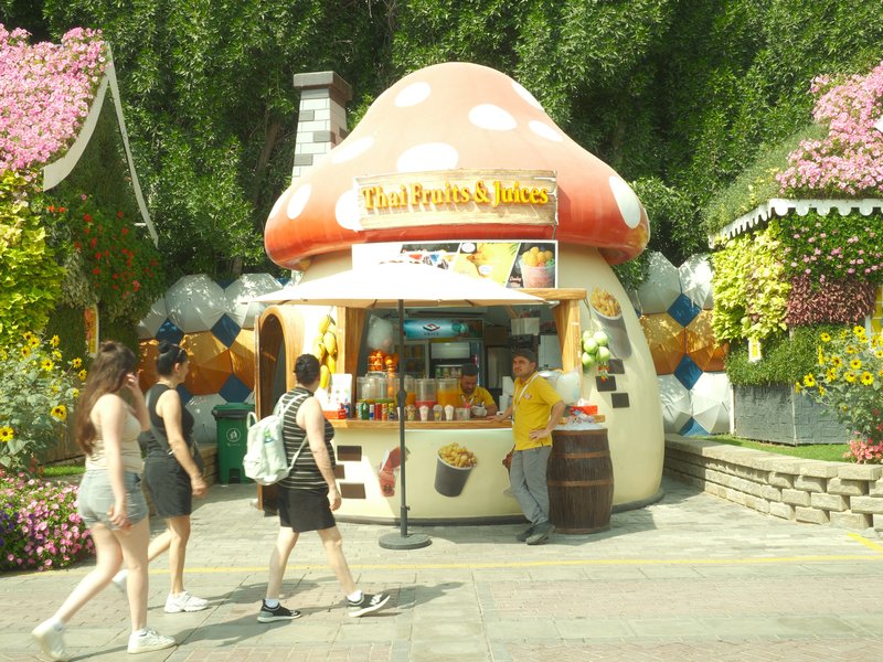 A whimsical mushroom-shaped kiosk surrounded by colorful flowers and people walking by.