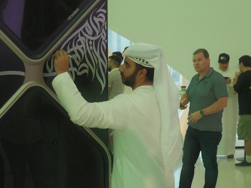 A man in traditional attire writing on a wall, with people observing in a modern indoor setting.
