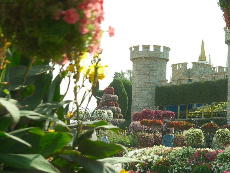 A colorful flower garden in front of a stone castle with towers and spires under bright sunlight.