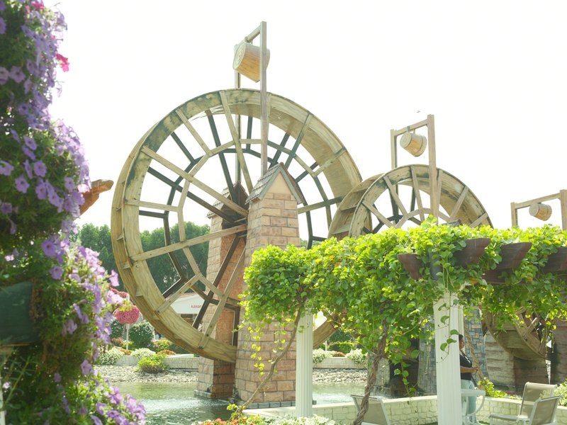 Two large water wheels surrounded by flowers and greenery in a bright outdoor setting.