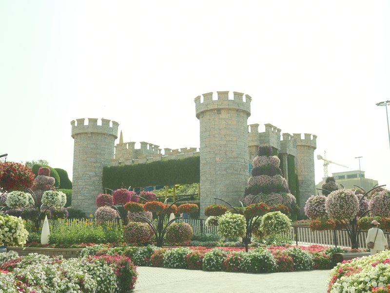A stone castle with towers surrounded by colorful flower beds in a bright outdoor setting.