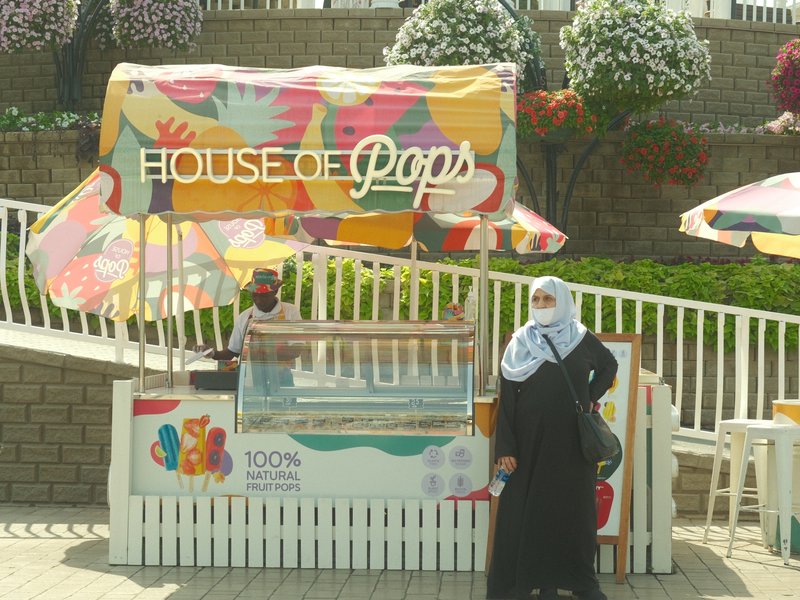 A colorful ice pop stand with a woman in a hijab standing beside it, set in a sunny outdoor environment.