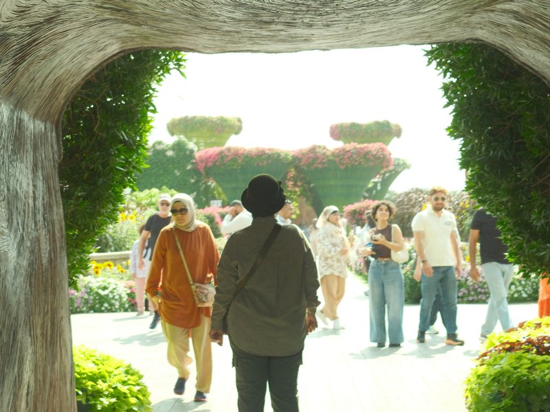 A person stands in silhouette at an archway, looking towards a bright garden filled with people.