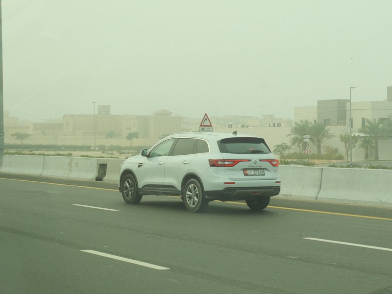 A white SUV driving on a hazy road with buildings in the background under overcast skies.