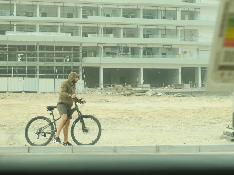 A person in casual attire riding a bicycle near a construction site with buildings in the background.