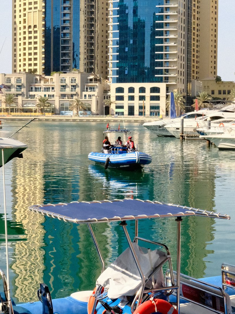 A small blue boat with four people on a calm water surface reflecting tall buildings.
