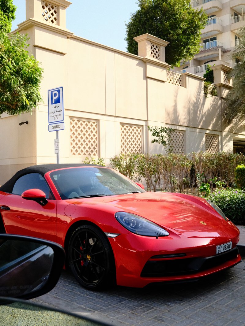 A vibrant red convertible sports car parked near a building with greenery and a parking sign.