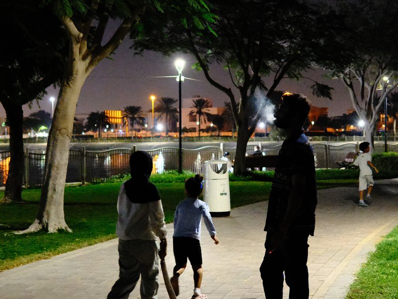 Silhouetted figures walking along a park path at night, with lights reflecting on water.