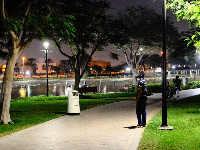A person standing on a path in a park at night, with trees and lights reflecting on a nearby water body.