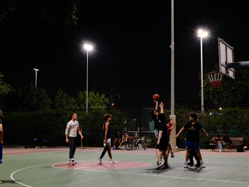 A group of people playing basketball at night on an outdoor court, with bright lights illuminating the scene.