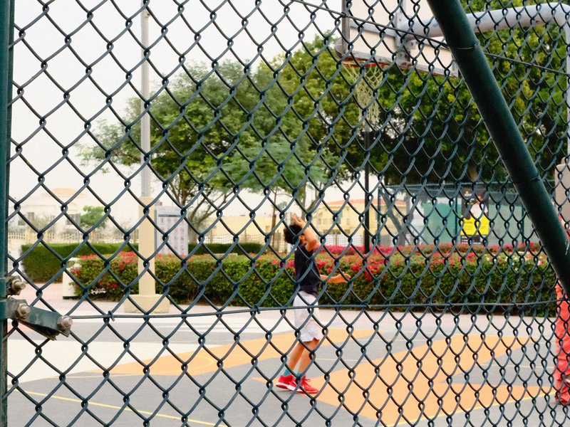 A young person playing basketball on an outdoor court, seen through a chain-link fence.