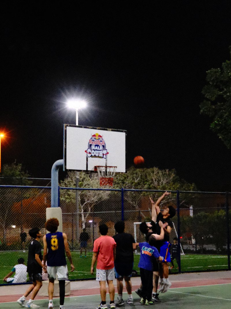 A group of young basketball players shooting hoops at night in a park with bright lights.