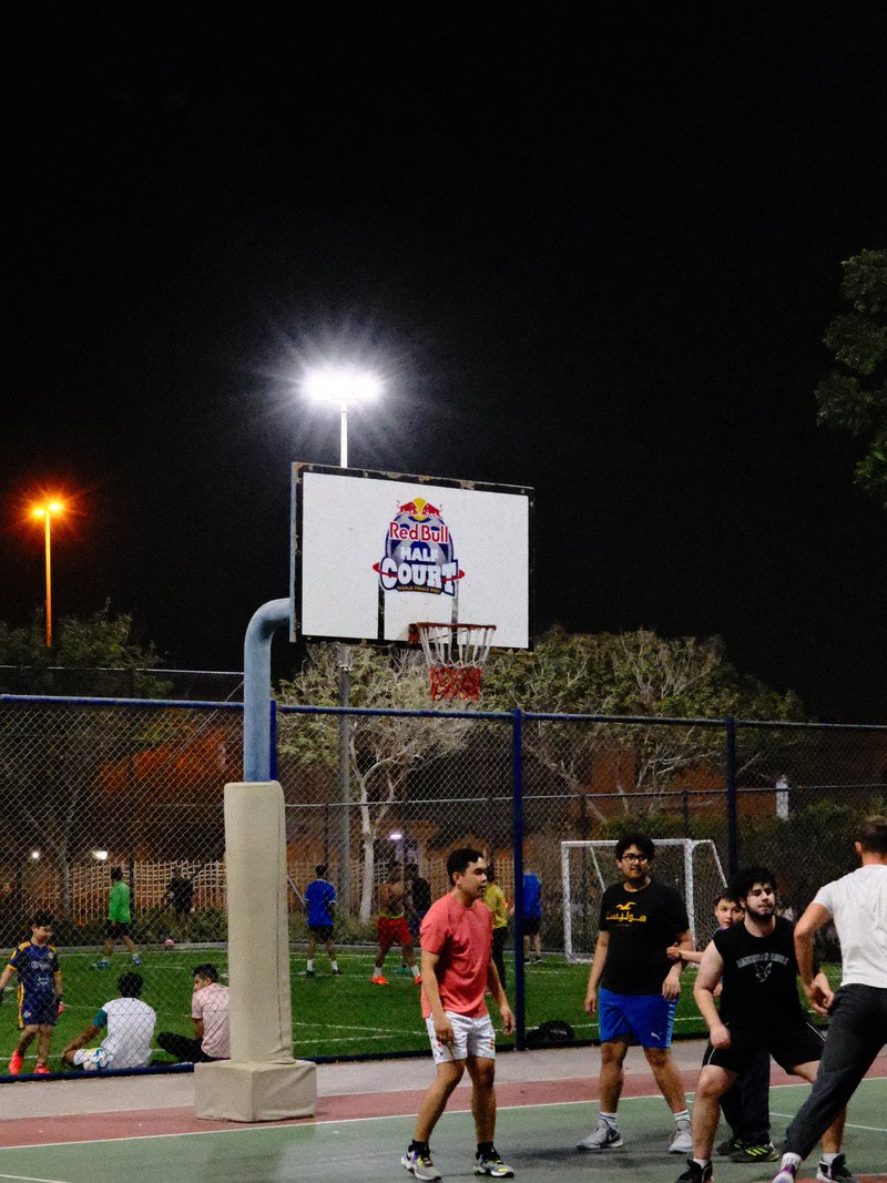 A nighttime basketball game with players on the court and a lit hoop in the background.