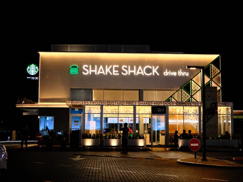 Exterior view of a Shake Shack restaurant at night, illuminated signage and nearby Starbucks.