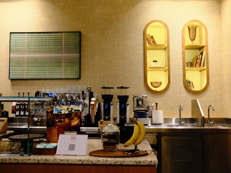 A cozy coffee shop counter with a coffee machine, bananas, and decorative shelves on the wall.