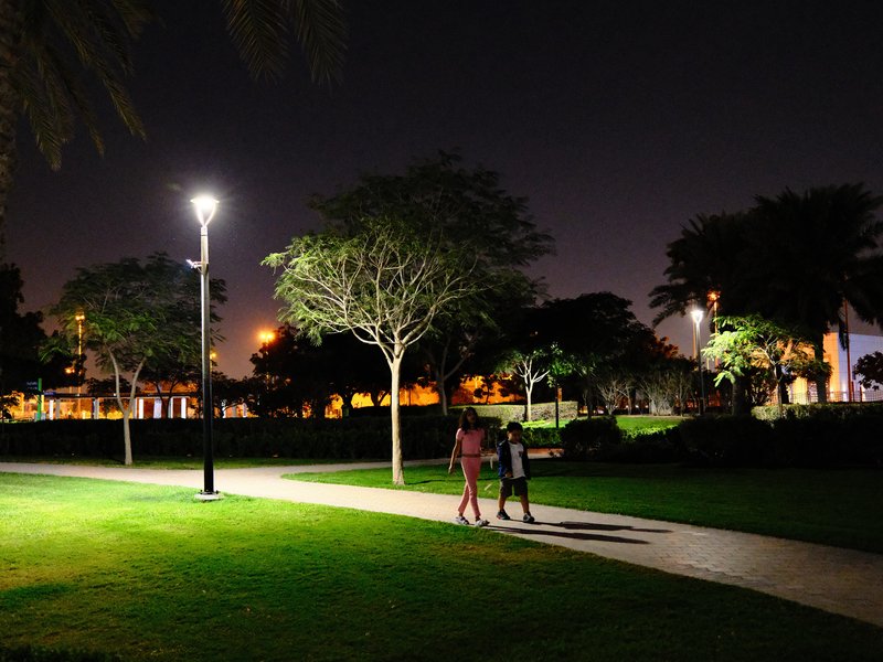 Two people walking on a path in a park at night, illuminated by streetlights and surrounded by trees.