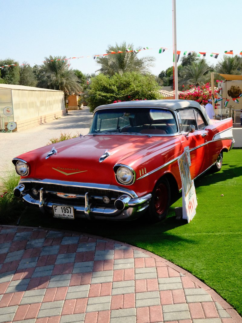 A classic red 1957 Chevrolet parked on green grass with palm trees in the background.