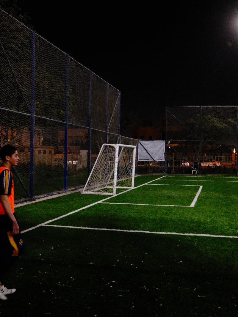 A young player stands near a soccer goal on a well-lit field at night.