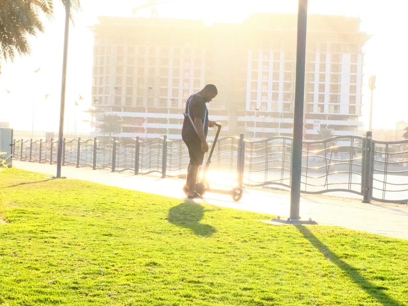 Silhouette of a person riding a scooter on a sunlit path with a building in the background.