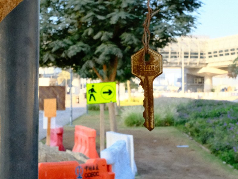 A close-up of a key hanging in the foreground with a blurred urban scene behind it, including signs and greenery.