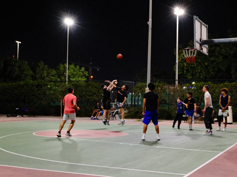A group of people playing basketball at night on an outdoor court, with bright lights illuminating the scene.