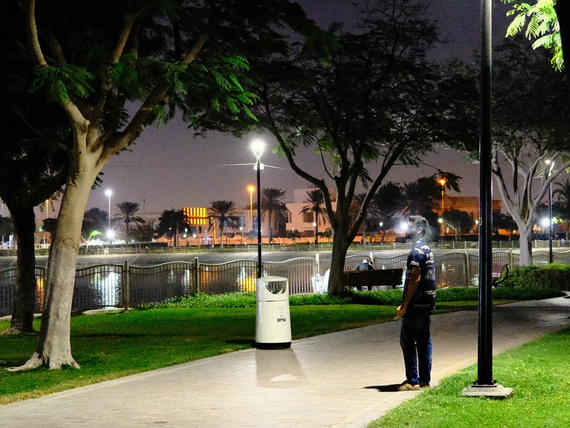 A person standing on a path by a river at night, surrounded by trees and streetlights.
