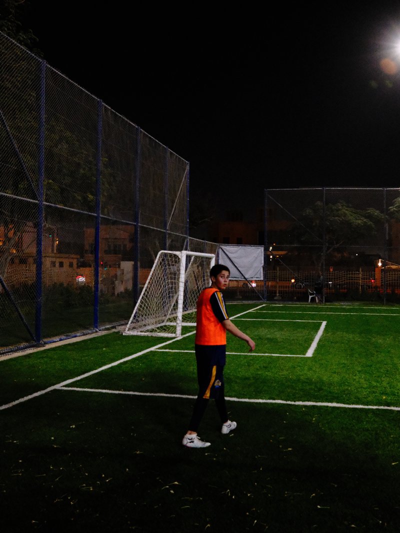 A young man in an orange shirt stands on a soccer field at night, with a goalpost in the background.