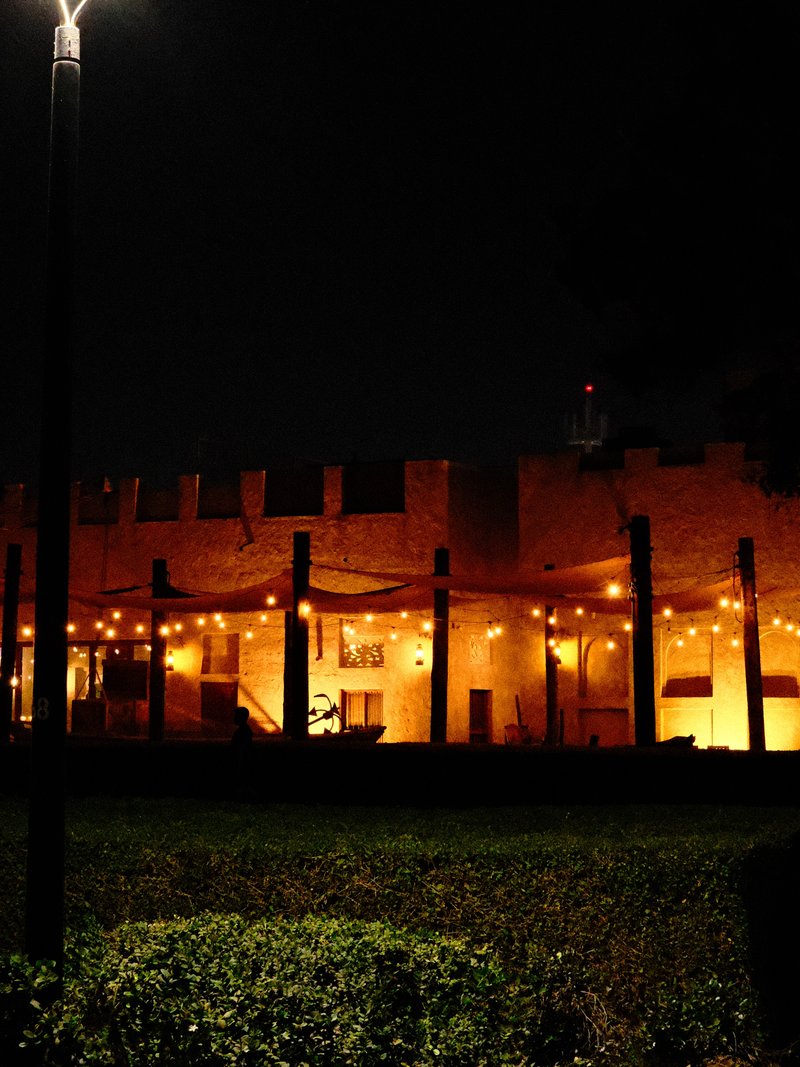 A historic building illuminated at night with string lights, surrounded by greenery.