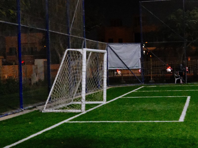 A soccer goal on a well-lit artificial turf field at night, with a blurred background of urban lights.