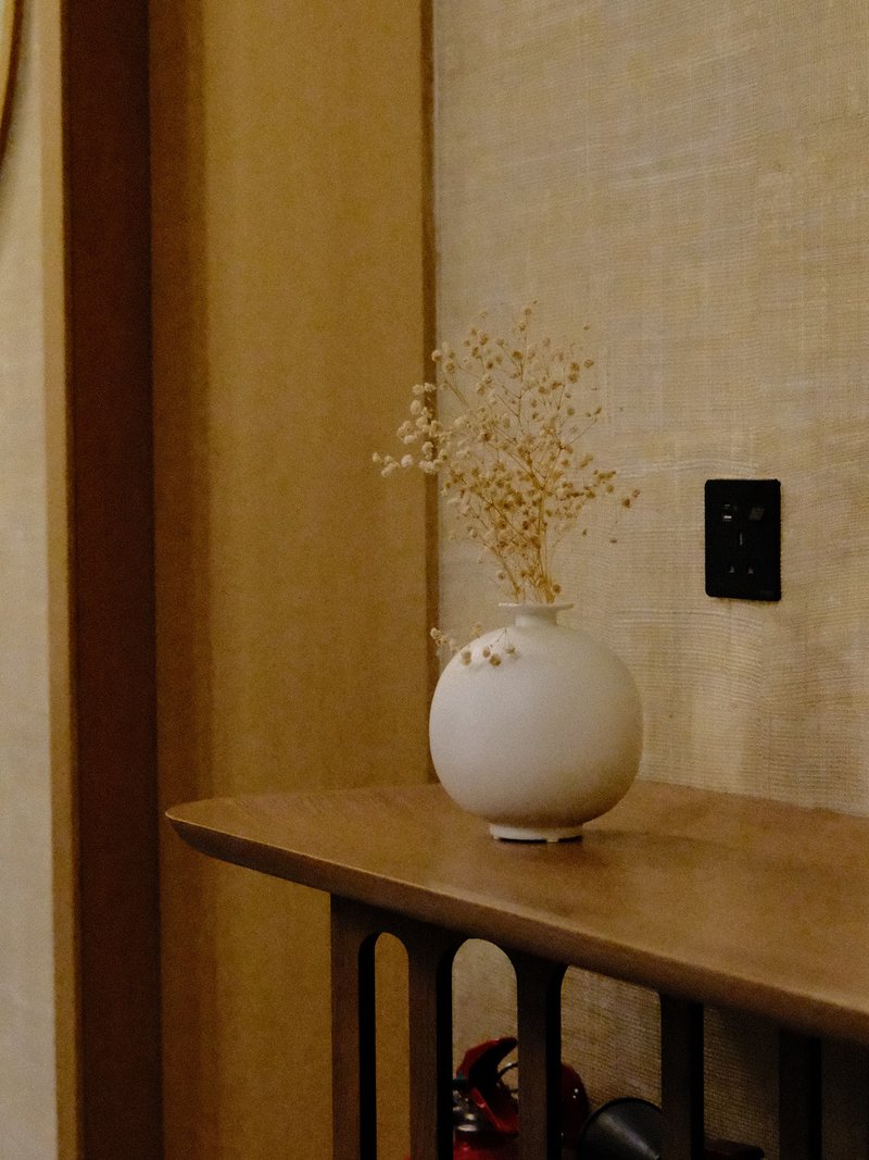 A minimalist vase with dried flowers on a wooden table against a textured beige wall.