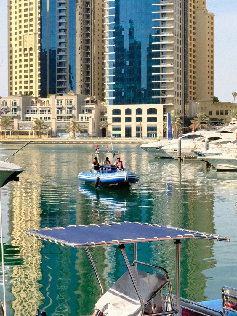 A blue boat with people on a calm water surface reflecting buildings and boats in a marina.