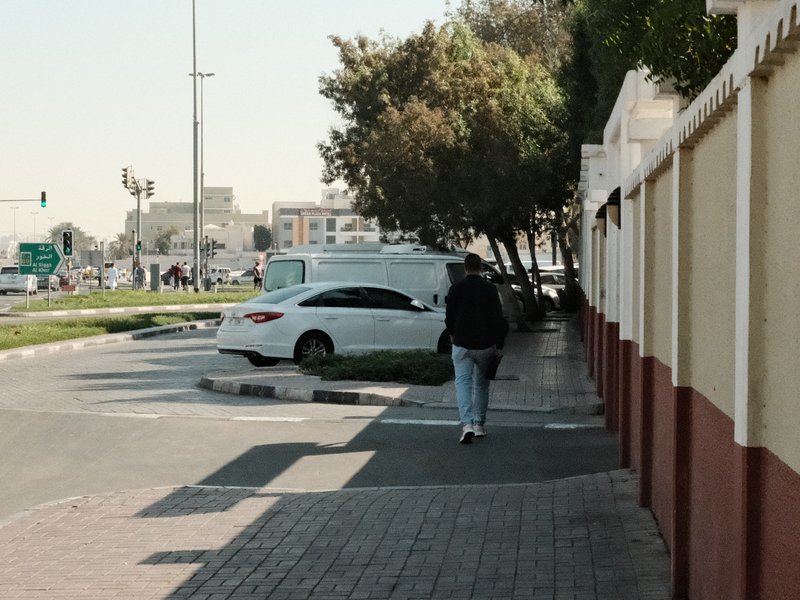 A person walking along a sidewalk next to a road with trees and a white car in the background.