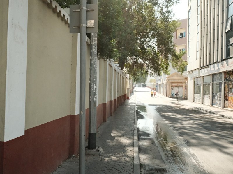 A quiet urban street with a wall on one side and buildings in the background under bright sunlight.