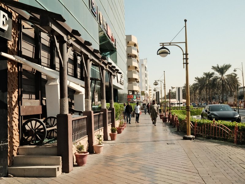 A sunny urban street scene with a wooden structure, palm trees, and pedestrians walking.